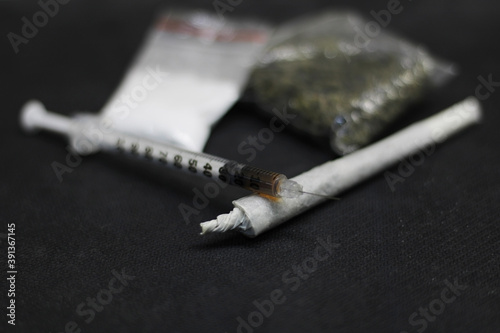 A syringe full of cocaine, placed on a cane of marjiuana or ganja, with a sachet full of cocaine, and a sachet full of ganja or marijuana placed in the background