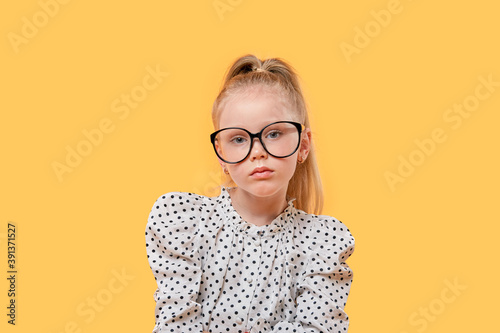 Portrait of a blonde Girl child with glasses for vision looking at the camera. Black-rimmed glasses. Yellow isolated background.