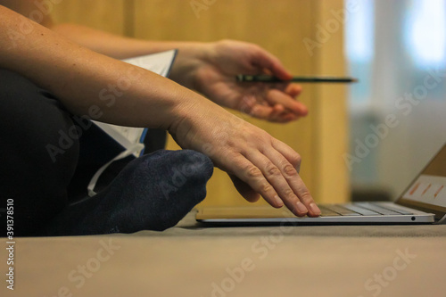 Remote study or work at the computer in a comfortable home environment, a girl sits on a gray sofa with a laptop and a diary and types text on the keyboard.