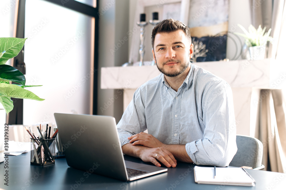Attractive freelancer guy in formal wear working remotely at home sitting at the table and using laptop, he looks at the camera. Distant work