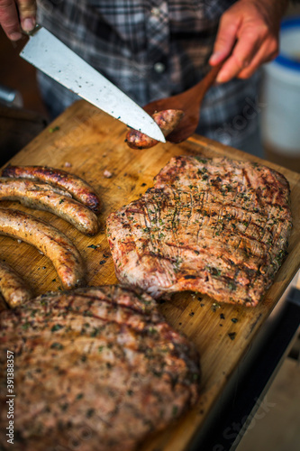 A man cuts sausage above a cutting board full of flank steak and brats photo