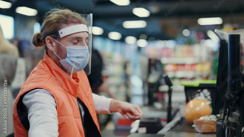 Bearded young cashier wearing respiratory mask face shield servicing young woman customer in supermarket grocery store during coronavirus quarantine.