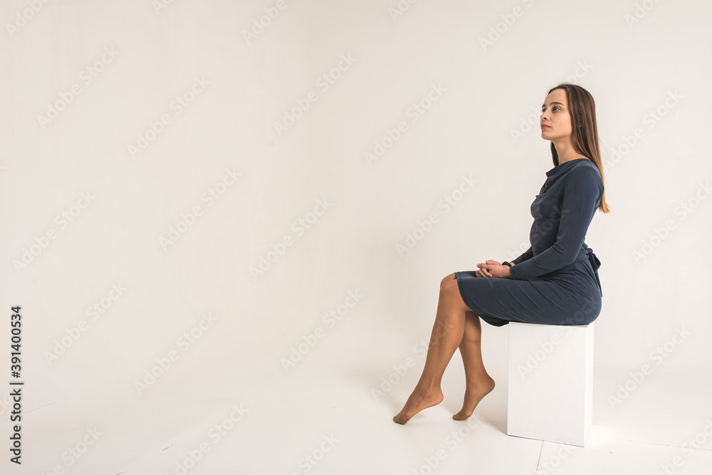 Young, pretty mixed race girl sitting on a white square, isolated on a white background, looking straight, thoughtful, serious
