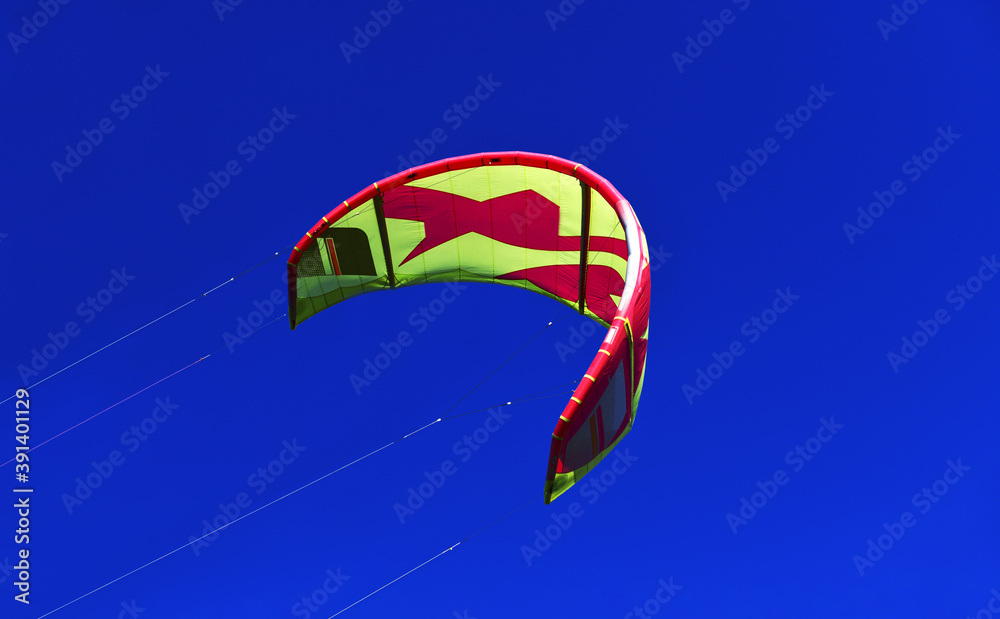 Bright kite, wind-curved kitesurfing parafoil flying in the blue sky. The slings are tight. Ripstop nylon, a flexible and durable curved wing. Flight over water. Bright kite, wind-curved kitesurfing parafoil flying in the blue sky. The slings are tight. Ripstop nylon, a flexible and durable curved wing. Flight over water.
