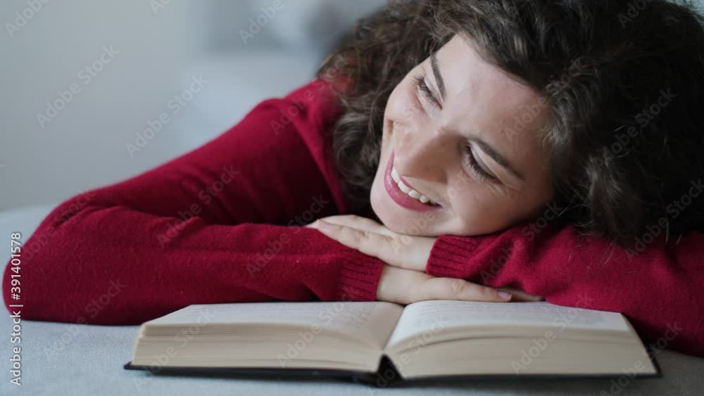 Happy young woman with red pullover resting her head on book. Pretty and cute millennial girl with curly brown hair looking in love in the distance. Slow-motion, close-up, natural daylight at home. Happy young woman with red pullover resting her head on book. Pretty and cute millennial girl with curly brown hair looking in love in the distance. Slow-motion, close-up, natural daylight at home.