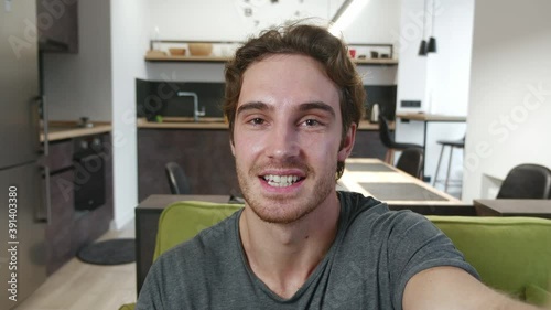 Smiling young man making selfie or video call while sitting at home on sofa. Portrait of male blogger doing a video chat from his apartment.