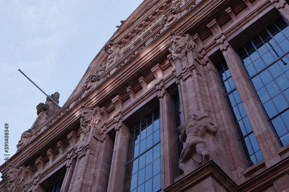 Obraz premium Low angle and exterior view of European classic facade with high glass windows, deep red stone wall and decorated sculpture against blue sky.