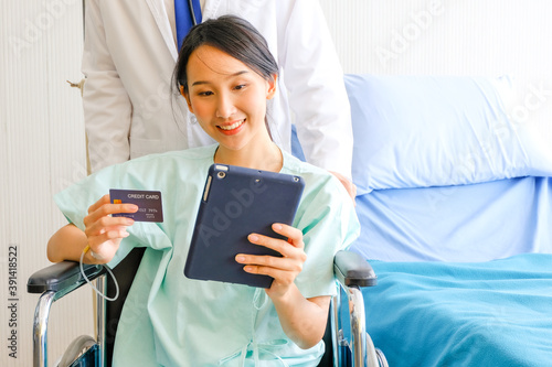 Happy Female Asian patient hold a credit card and Tablet while sitting in wheelchair in hospital with doctor. Patient smiling no worry about the payment. Healthcare, medical concept.
