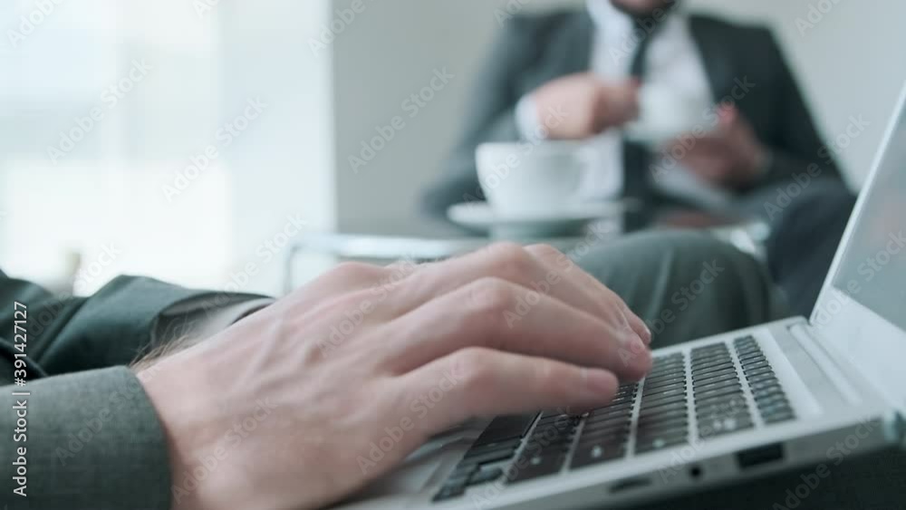Unrecognizable man sitting at low table in office hall typing something on laptop while having coffee break with his colleague Unrecognizable man sitting at low table in office hall typing something on laptop while having coffee break with his colleague