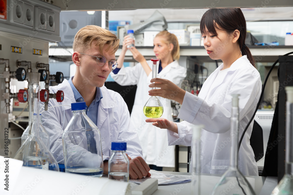 Fototapeta premium Positive Chinese female working with reagent in test tube during chemical experiment with male fellow student in university lab. Focus on male