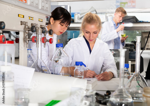 Two female students of Faculty of Chemistry performing experiments in university laboratory  recording results in workbook