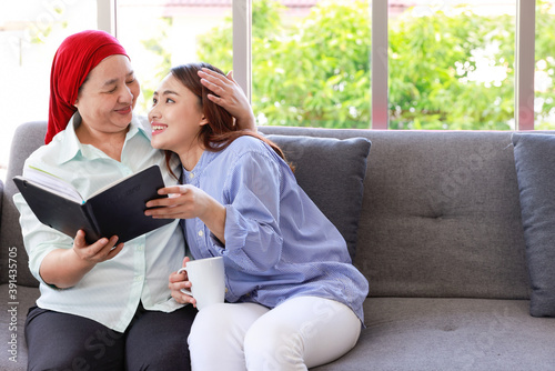 A senior woman with cancer wearing a headscarf relaxes at home with her adult daughter and reading a book with smiling.The women are full of hope for the future..