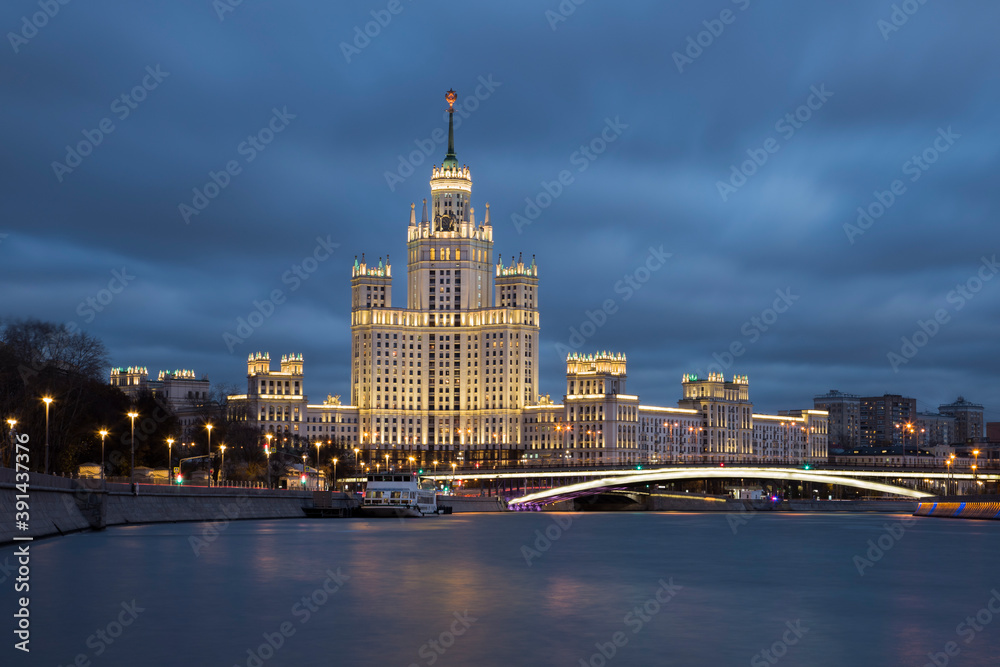 Night cityscape of brightly lighted empire style skyscraper on a riverbank. Bright bridge in front of the building. Blurred clouds on a blue-hour sky. Blurred reflections in the water. No people