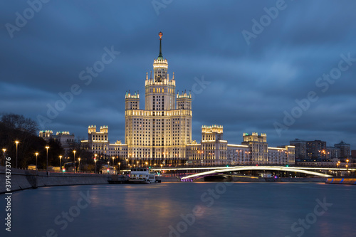 Night cityscape of brightly lighted empire style skyscraper on a riverbank. Bright bridge in front of the building. Blurred clouds on a blue-hour sky. Blurred reflections in the water. No people
