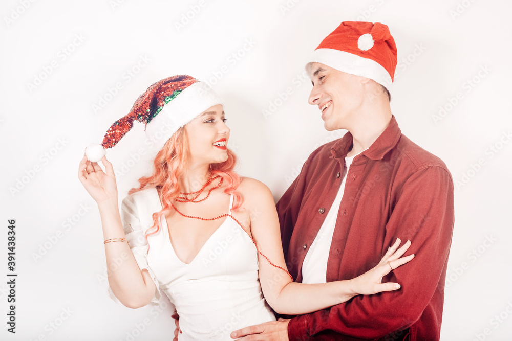 Obraz premium Couple on a white background in santa hats. The girl and the guy are smiling, new year, christmas. Woman in a beautiful white dress and pink hair, in accessories
