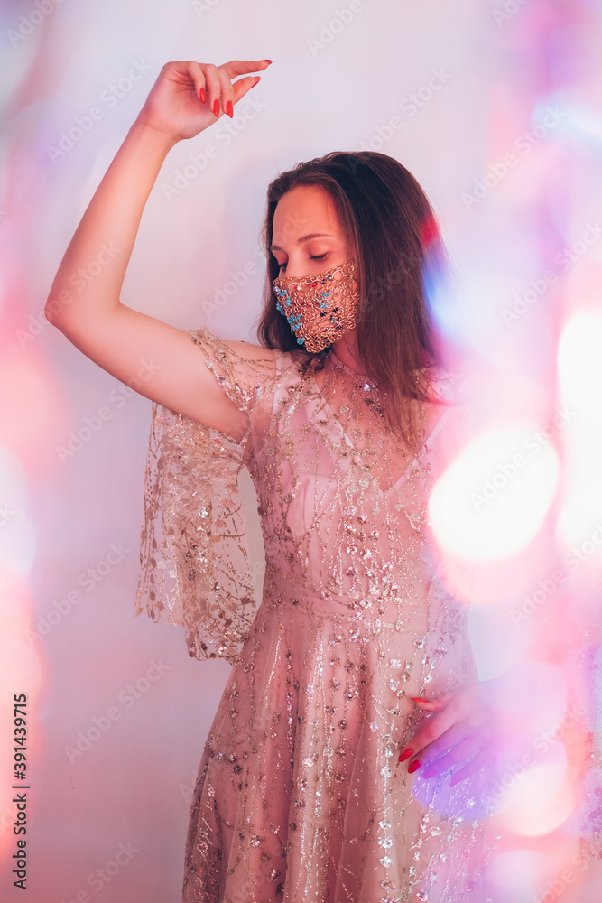 Naklejka premium Christmas festive look. Pandemic fashion. Ethnic style. Beautiful woman in east glossy dress designer golden chain crystal beads face mask posing in pastel neon light blur lens flare.