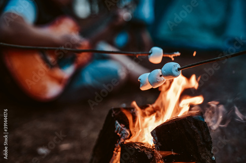 Romantic and atmospheric photography of marshallow on sticks its toasting at the stake in blurred background of guitarist and forest.