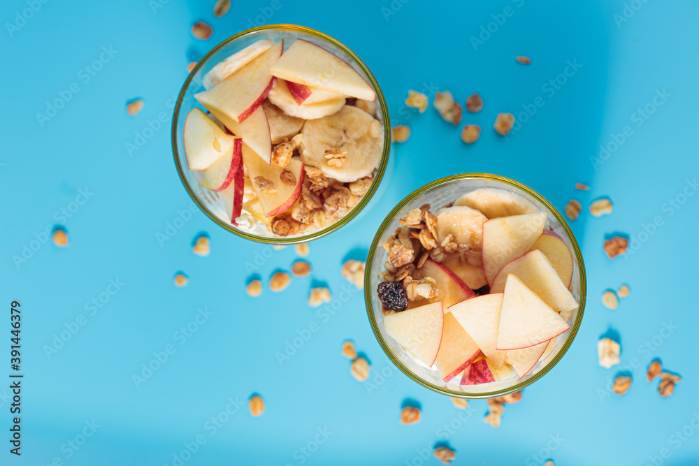 Obraz Prawidłowe odżywianie, śniadanie. Welness - granola z jogurtem i owocami: bananem i jabłkiem, w szklanych kubkach na niebieskim tle, widok z góry.