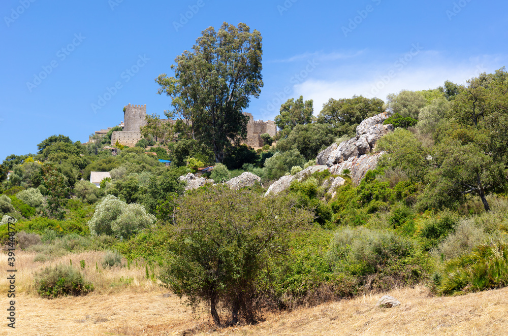 The castle of Castellar is surrounded by a small mountain village in Andalusia on the edge of a reservoir. It's a sunny summer day with blue skies and clouds.