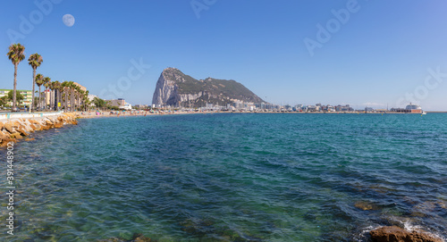 View from mainland Spain to the British territory of Gibraltar on a summer day with a blue sea. On the left is the access road with palm trees. A cruise ship is on the right.