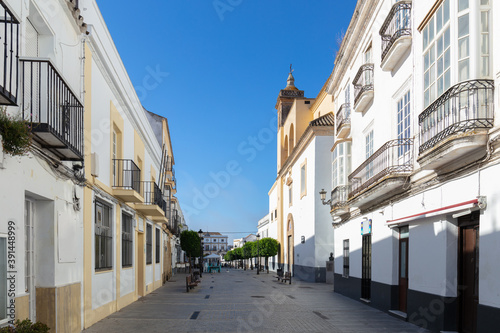 A street in the historic mountain village of Medina-Sidonia in Andalusia in southern Spain with a church and green trees. The houses are all painted white. It's early morning with sunshine.