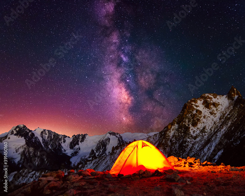Camping in the highlands under the night sky with stars; bright orange tent in the mountains on the background of majestic Milky Way