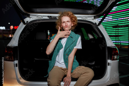 A cool-looking redhead young woman with freckles looking into a camera sitting and posing inside of an opened car trunk outside on a parking site with a led screen behind