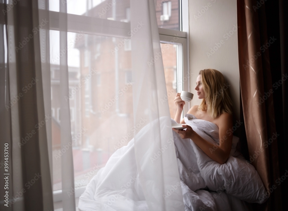 woman wrapped in a blanket sits on the windowsill by a large window woman wrapped in a blanket sits on the windowsill by a large window