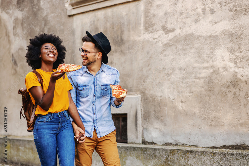 Full length of happy multiracial couple walking outdoors, holding hands and eating pizza.