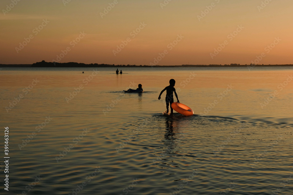 Orange inflatable circle and baby silhouette on sunset background. The child walks in shallow water. Copy space.
