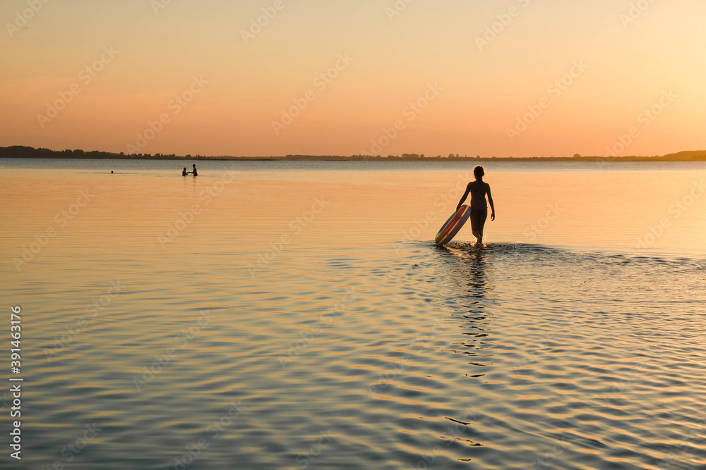 Girl silhouette on a background of sunset. The girl walks in shallow water with an inflatable circle. Copy space. 