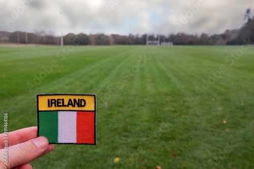 Badge with sign Ireland and Irish National flag in focus, Training ground for Irish sports out of focus. Concept hurling, camogie, rugby, gaelic football. Nobody photo