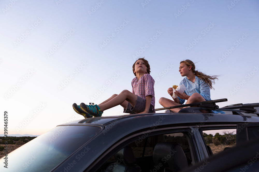 Teenage girl and her younger brother sitting atop their SUV at sundown. Teenage girl and her younger brother sitting atop their SUV at sundown.