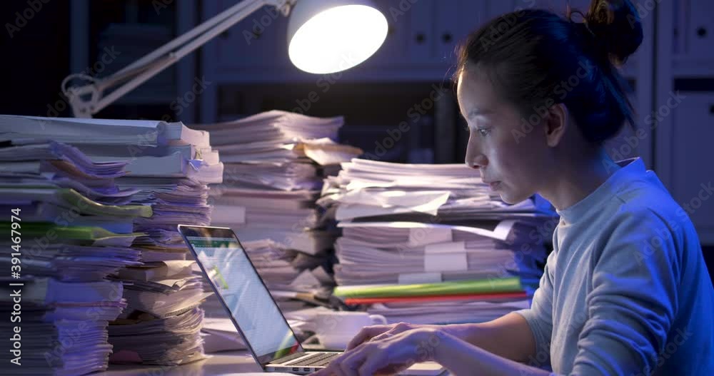 Asian business woman is sitting at desk covered with stack of paperwork and looking at paper report, typing on notebook. Alone girl checking document while working hard at late night. Side view shot.