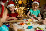 Mother and little kids in red hats cooking gingerbread cookies and playing. Beautiful living room with lights and Christmas tree, table with lantern. Happy family celebrating holiday together.