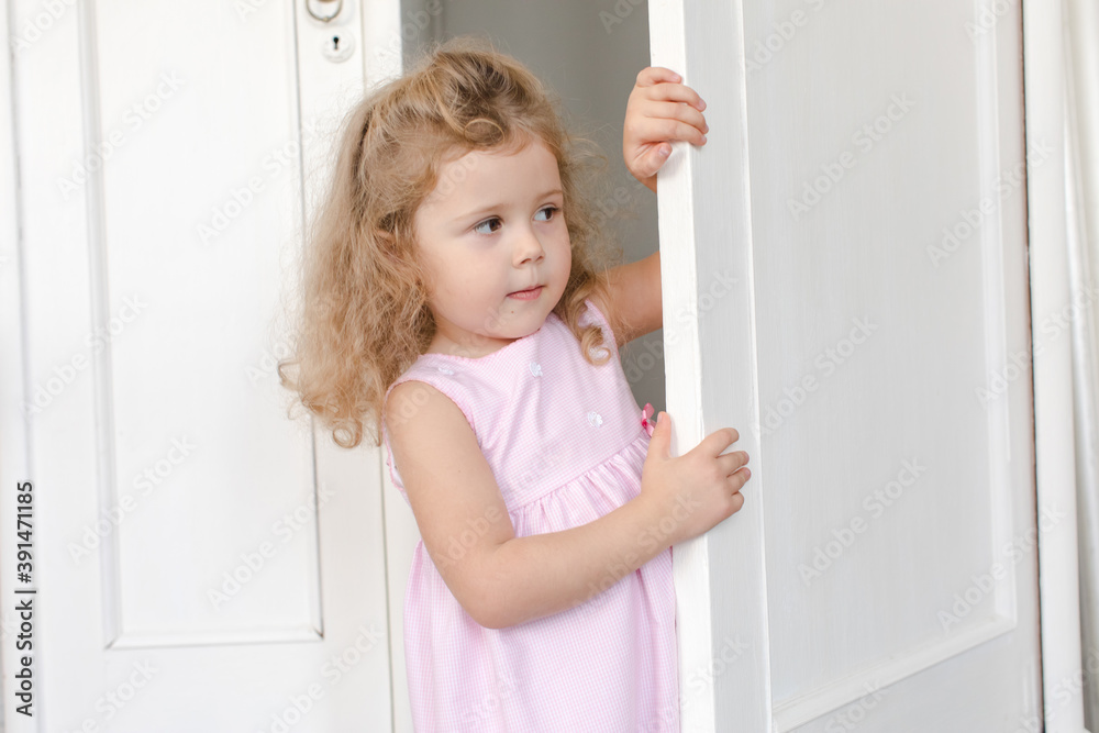 Curious little child in dress peeping out of door while playing hide and seek in flat Curious little child in dress peeping out of door while playing hide and seek in flat