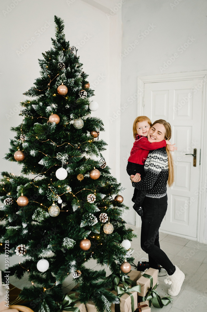 Merry Christmas and Happy Holidays. Cheerful mom hugging cute baby daughter girl near Christmas tree. Mother and little child having fun and playing together at home.