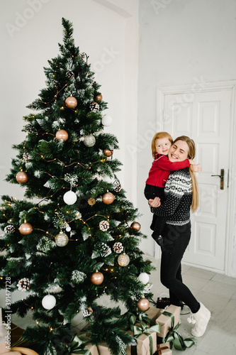 Merry Christmas and Happy Holidays. Cheerful mom hugging cute baby daughter girl near Christmas tree. Mother and little child having fun and playing together at home.