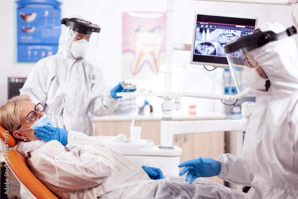 Fototapeta premium Senior patient presenting teeth pain wearing hazmat suit agains coronavirus at dentist. Elderly woman in protective uniform during medical examination in dental clinic.