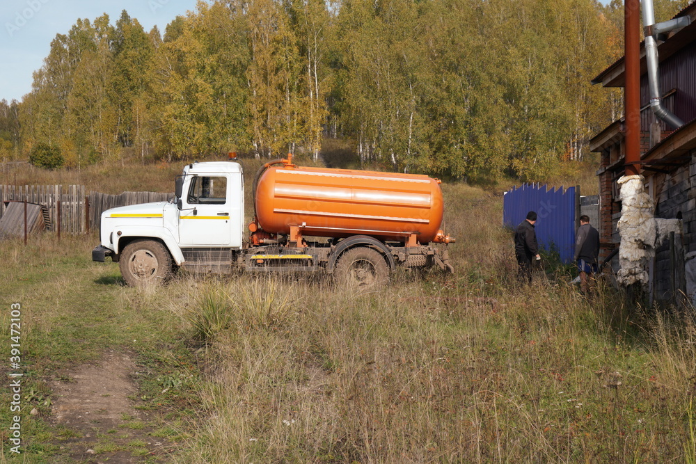 A cesspool machine and two men are standing near a village building amid an autumn birch grove.
