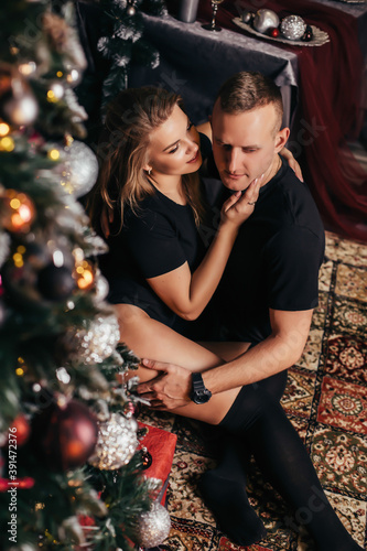 Attractive couple at Christmas at home near a Christmas tree hugging each other. New Year's photo of a guy and a girl in a New Year's interior, a cozy decorated house. © Дмитрий Ткачук