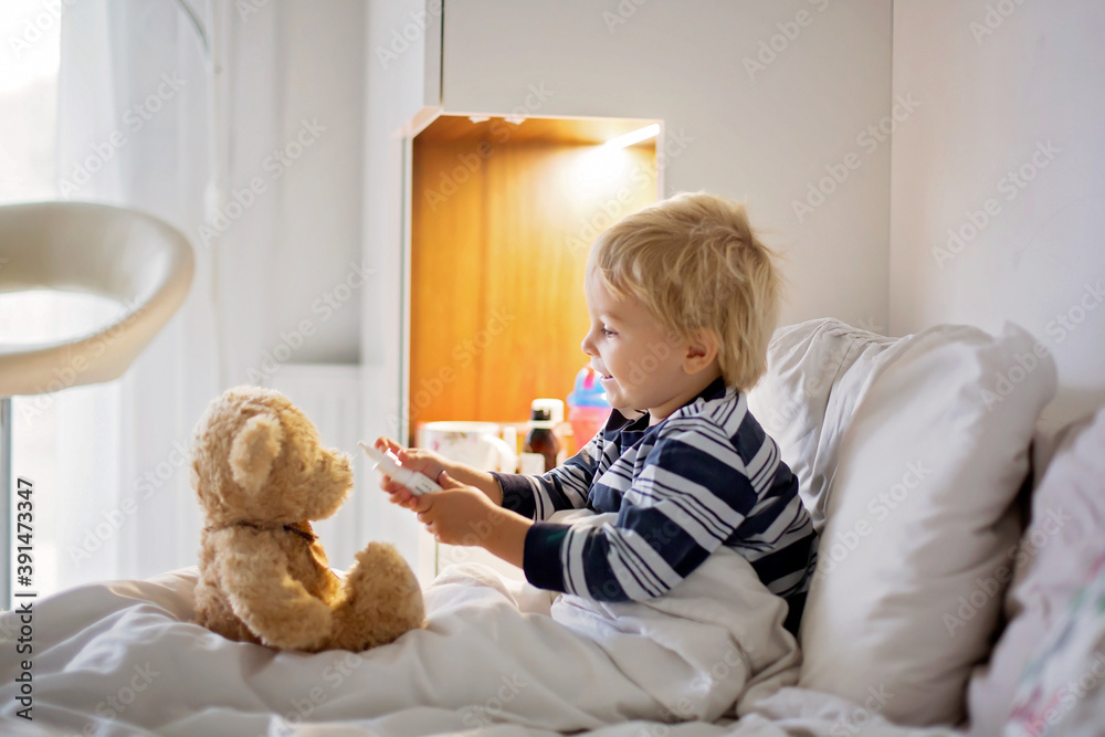Little toddler blond child, playing with teddy bear in bed, while being sick, checking his temperature Little toddler blond child, playing with teddy bear in bed, while being sick, checking his temperature