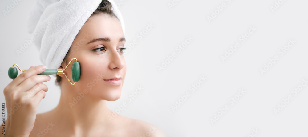 Beautiful young woman with perfect skin wearing towel on head using a jade face roller with natural quartz stones. Place for text