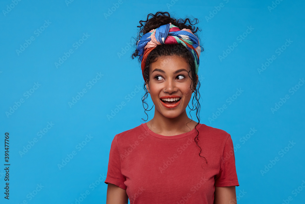 Indoor photo of young lovely dark haired curly female with nose piercing looking excitedly aside with wide smile, standing over blue background with hands down Indoor photo of young lovely dark haired curly female with nose piercing looking excitedly aside with wide smile, standing over blue background with hands down