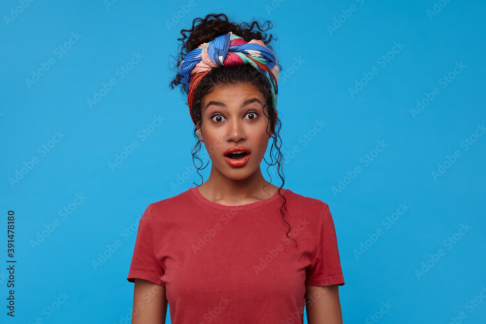 Amazed young pretty brunette curly lady with gathered hair looking surprisedly at camera with wide eyes and mouth opened, standing over blue background Amazed young pretty brunette curly lady with gathered hair looking surprisedly at camera with wide eyes and mouth opened, standing over blue background