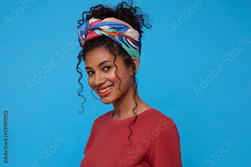 Studio photo of young lovely brown-eyed curly dressed in burgundy t-shirt and colored headband looking gladly at camera with charming smile, isolated over blue background