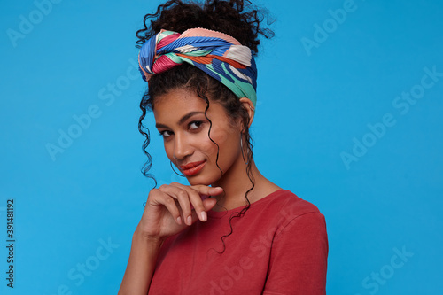 Pensive young pretty brown-eyed curly female with casual hairstyle leaning her chin on raised hand and smiling slightly while looking gently at camera, posing over blue background