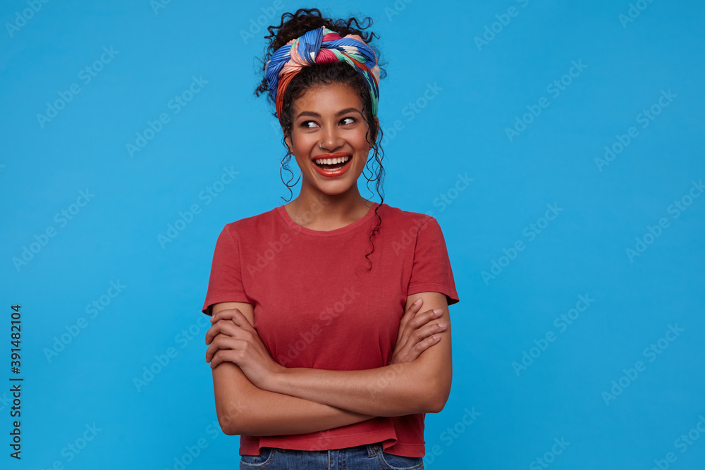 Glad young attractive dark haired curly female with festive makeup keeping hands folded while looking cheerfully aside and smiling widely, isolated over blue background Glad young attractive dark haired curly female with festive makeup keeping hands folded while looking cheerfully aside and smiling widely, isolated over blue background