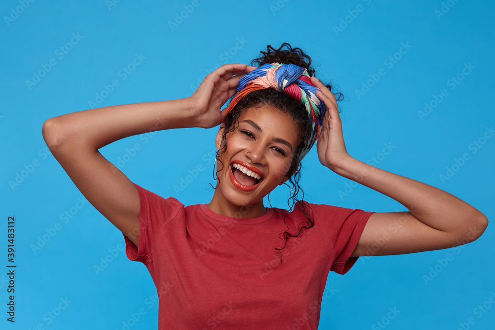 Portrait of young lovely dark haired curly female with festive makeup laughing happily and raising emotionally hands to her head, isolated over blue background Portrait of young lovely dark haired curly female with festive makeup laughing happily and raising emotionally hands to her head, isolated over blue background