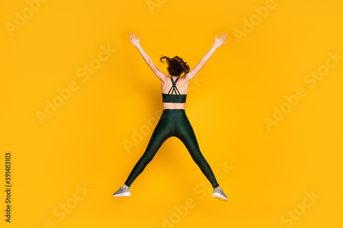 Full size behind view photo of sportive lady jump high up spread hands wear sports suit sneakers isolated yellow color background